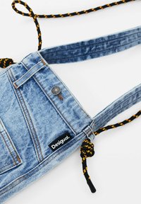 Denim-Crossbody-Tasche in hellblauer Farbe mit einem Reißverschlussfach, einem Logo-Tag und einem schwarzen Tragegurt mit gelben Akzenten, die eine strukturierte Oberfläche zeigt.