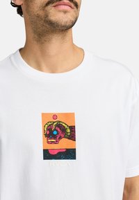 Hand holding a pink skull with blue eyes within an orange and black rectangular graphic on a white T-shirt.