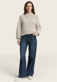 Pull en ribbed beige clair avec un col montant, associé à un jean bleu foncé à jambes larges et des baskets blanches. Design simple et coupe décontractée.