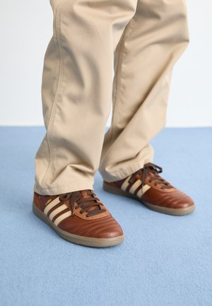 Bruine leren sneakers met crème strepen, een getextureerde neus en bruine veters; gedragen met beige, loszittende broek op een lichtblauwe ondergrond.