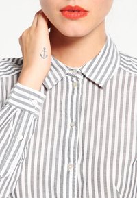 Chemise à rayures gris et blanc. Dotée d'un col classique et de manches retroussées avec des accents de boutons. Matériau en coton, texture légère.