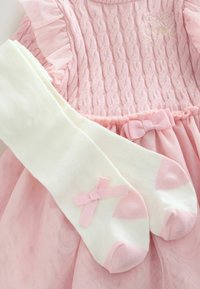 Vaaleanpunainen palmikkoneulemekko, jossa on röyhelöinen tyllihame ja rusetti, yhdistetty valkoisiin polvisukkiin, joissa on vaaleanpunaiset kärjet ja pienet vaaleanpunaiset rusete.