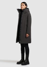 Dlhý tmavošedý kabát s kapucňou, ktorý má predný zips, bočné vrecká a elegantnú, hladkú textúru. Kombinovaný s čiernymi legínami a čižmami.