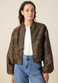 Veste bomber à imprimé léopard en marron et noir, avec fermeture éclair à l'avant, col côtelé et manches surdimensionnées. Associée à un jean en denim bleu clair.
