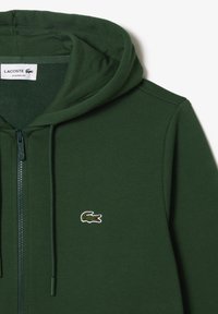 Sweat à capuche zippé vert foncé avec cordons, avec un patch logo crocodile Lacoste sur la poitrine gauche et une étiquette "Lacoste Classic Fit" à l'intérieur.