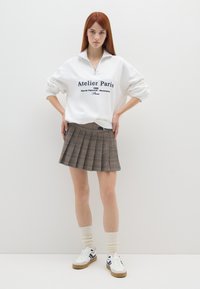 Pullover bianco in pile con zip e testo blu navy, abbinato a una gonna a pieghe scozzese marrone. Sneakers bianche con accenti blu navy e calze crema.