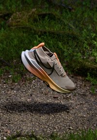 Beige joggesko med en pustende overdel, oransje detaljer, en polstret såle og en svart swoosh-logo. Designet for komfort og ytelse.