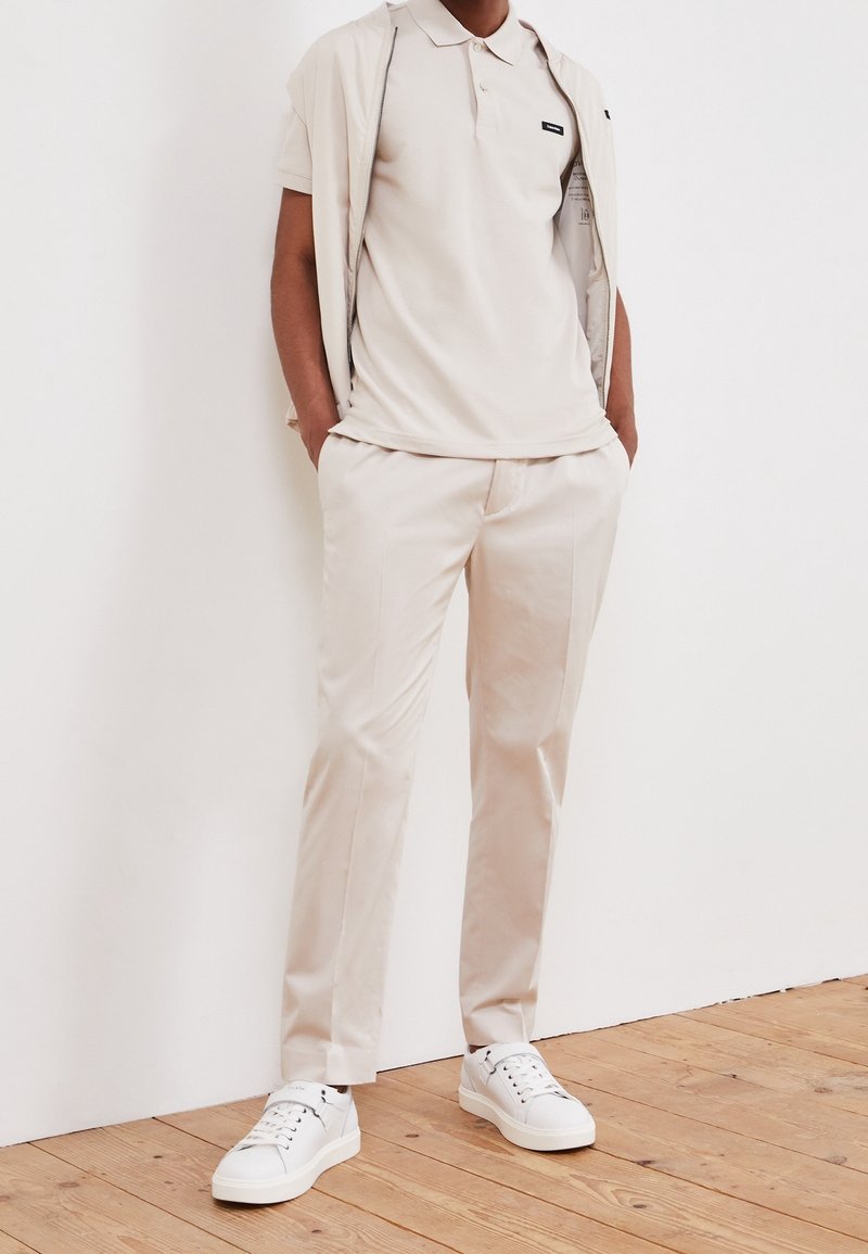 Homme vêtu d'un polo beige, d'un pantalon assorti et de baskets blanches, debout les mains dans les poches contre un mur blanc.