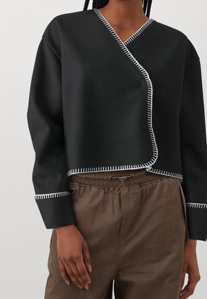 Close-up van een persoon die een zwart langärmelig shirt met witte stiknaad en bruine broek met trekkoord draagt.