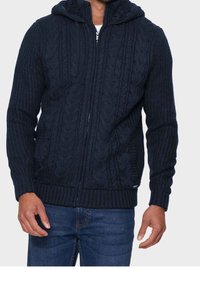 Marinblå stickad zip-up hoodie med en texturerad kabelmönster, ribbade muddar och fåll, samt en foderhuva. Har en silverdragkedjedetalj.