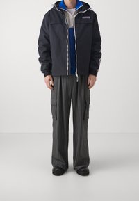 Czarna kurtka na zamek z kapturem, białe sznurki; pod spodem warstwowa szara i niebieska bluza. Luźne czarno-szare spodnie w paski z kieszeniami typu cargo.