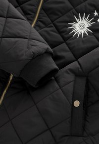 Fekete steppelt dzseki arany cipzárral, bordázott mandzsettákkal és hímzett nap motívummal csillagokkal. Puha tapintású és tartós anyagból készült.