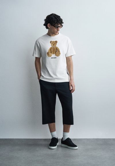 Homme portant des lunettes de soleil, un t-shirt blanc avec un motif d'ours en peluche, un pantalon noir court, des chaussettes blanches et des baskets noires, debout contre un mur uni.