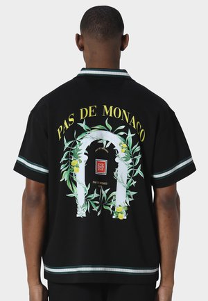 Rückansicht einer Person, die ein schwarzes Shirt mit einem laubumrankten Bogenmuster, gelben Zitronen und gelbem Text mit der Aufschrift "PAS DE MONACO" trägt.