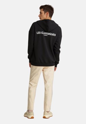 Black hoodie with white text "LES CLASSIQUES" on the back, paired with light-colored pants and beige sneakers. Smooth fabric, relaxed fit.