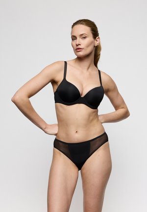 Woman posing in black underwire bra with double straps and matching sheer panel bikini panties against plain gray background.