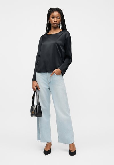 Γυναίκα με μακριά πλεξούδα που φοράει μαύρο σατέν μπλουζάκι με μακριά μανίκια, ανοιχτό μπλε τζιν παντελόνι με φαρδιά μπατζάκια, μαύρες γόβες και κρατάει μικρή μαύρη τσάντα χειρός.