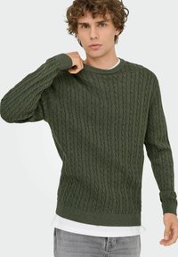 Pull en maille torsadée vert foncé avec un col rond côtelé et des manches longues, superposé sur une chemise blanche, comprenant des motifs texturés en relief.