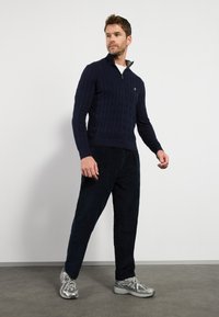 Marineblauw kabelgebreid trui met een halve rits, gedragen met een donkere corduroy broek en zilveren sportschoenen, tegen een neutrale achtergrond.