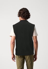 Black sleeveless knit sweater with a ribbed hem, worn over a light-colored t-shirt, featuring a minimalist design and textured surface.