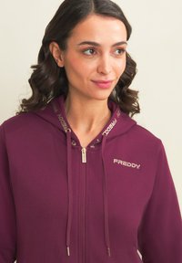 Felpa con zip color vino, dotata di cappuccio, con dettagli in oro e un logo. Realizzata in morbido tessuto con una superficie liscia.