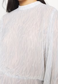 Femme portant une blouse transparente à manches longues avec un léger motif zèbre et un col haut.