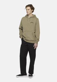 Felpa verde oliva con tasca frontale e logo nero, abbinata a pantaloni neri e sneaker nere. Tessuto morbido, vestibilità rilassata.