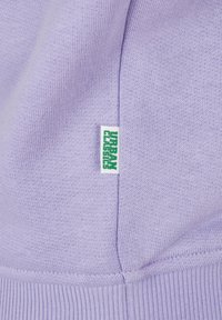 Lila pulover s mekim teksturom, rebrastim rubom i malom bijelom etiketom s zelenim natpisom "URBAN CLASSICS" sa strane.
