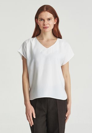Junge Frau mit glattem roten Haar, die eine weiße kurzärmelige V-Ausschnitt-Bluse und dunkle Hose trägt, steht vor einem einfachen Hintergrund.