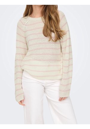 Femme portant un pull en maille crème avec des rayures horizontales rose clair et un pantalon blanc, avec de longs cheveux bruns lâches sur une épaule.