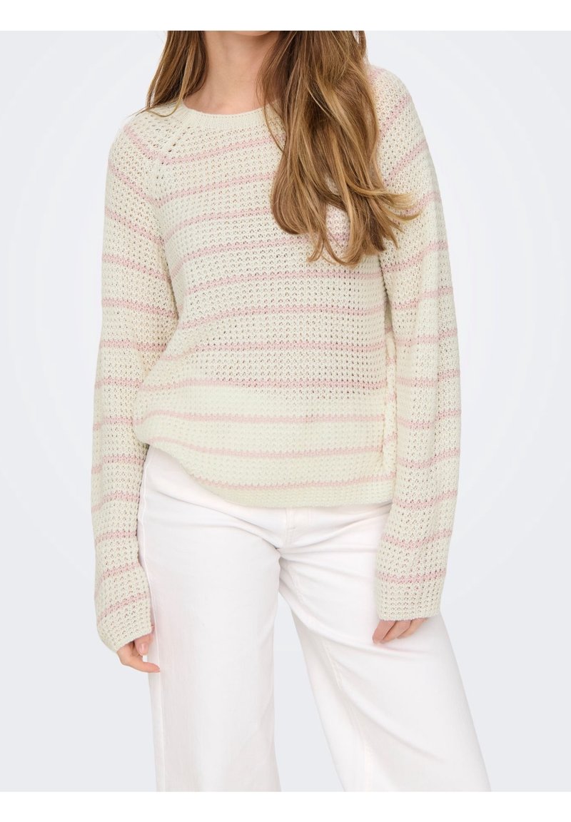 Femme portant un pull en maille crème avec des rayures horizontales rose clair et un pantalon blanc, avec de longs cheveux bruns lâches sur une épaule.