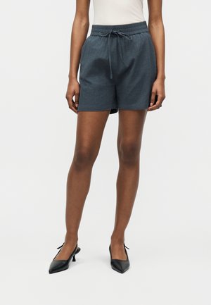 Ženska obleka temnosivih kratkih hlač z elastičnim pasom in vrvico ter črne salonarje z zabodnimi konicami, ki stoji ob svetli enobarvni podlagi.