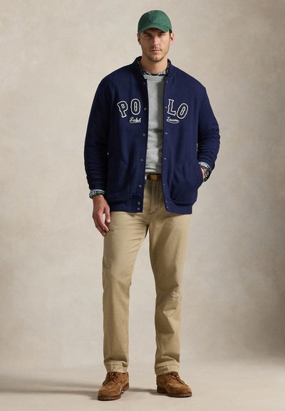 Άνδρας με μπλε ζακέτα varsity με γράμματα "POLO", πράσινο καπέλο, μπεζ παντελόνι, καφέ παπούτσια και φορεμένο πουκάμισο με στρώσεις, στέκεται μπροστά σε απλό φόντο.