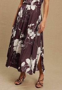 Langes lilafarbenes Kleid mit weißem Blumenmuster, das eine geraffte Taille und seitliche Schlitze aufweist. Begleitet von schwarzen Riemchen-Heels.