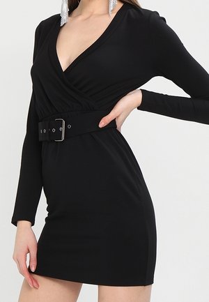 Frau trägt ein enganliegendes schwarzes Kleid mit langen Ärmeln, tiefem V-Ausschnitt und breitem Taillengürtel, posiert mit einer Hand in der Hüfte.