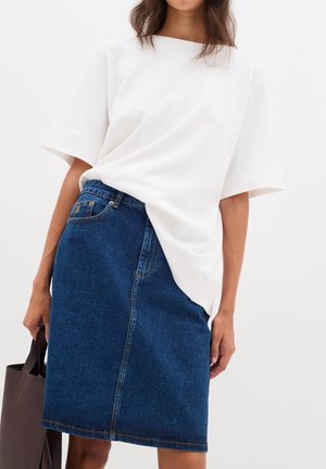 Vrouw draagt een wit los T-shirt in een knielange blauwe spijkerrok en houdt een donkerbruine handtas vast.