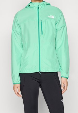 Veste à capuche zippée vert clair avec poche poitrine, portée par une personne en pantalon noir avec un logo visible sur la cuisse gauche.