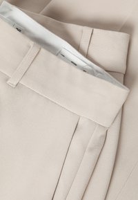 Close-up van de tailleband van beige op maat gemaakte broek met riemlussen en een verstelbare binnenhaak-en-oogsluiting.