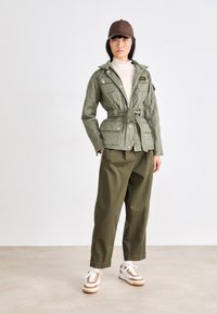 Olijfgroene gewatteerde jas met een ceintuur in de taille, gouden hardware, beige coltrui, olijfgroene broek en witte sneakers met bruine accenten.