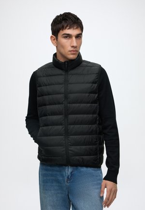 Jeune homme portant un gilet matelassé noir sans manches par-dessus une chemise noire à manches longues et un jean bleu, debout avec une main dans la poche.
