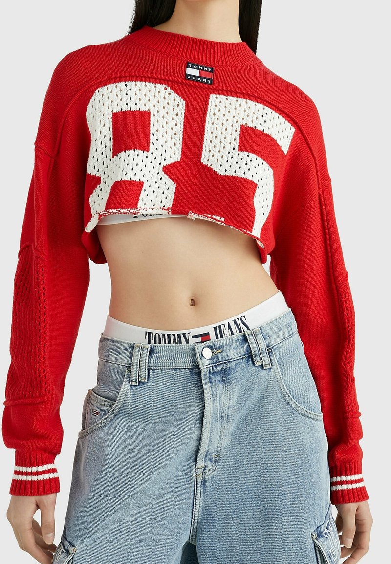 Červený zkrácený svetr s bílým pleteným číslem "85", žebrovaným výstřihem a manžetami, spárovaný se světle modrými džíny s vysokým pasem a elastickým pasem značky.
