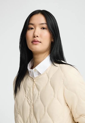 Joven mujer con cabello liso y negro, vistiendo una camisa blanca de cuello y una chaqueta acolchada color beige claro, posando frente a un fondo liso.