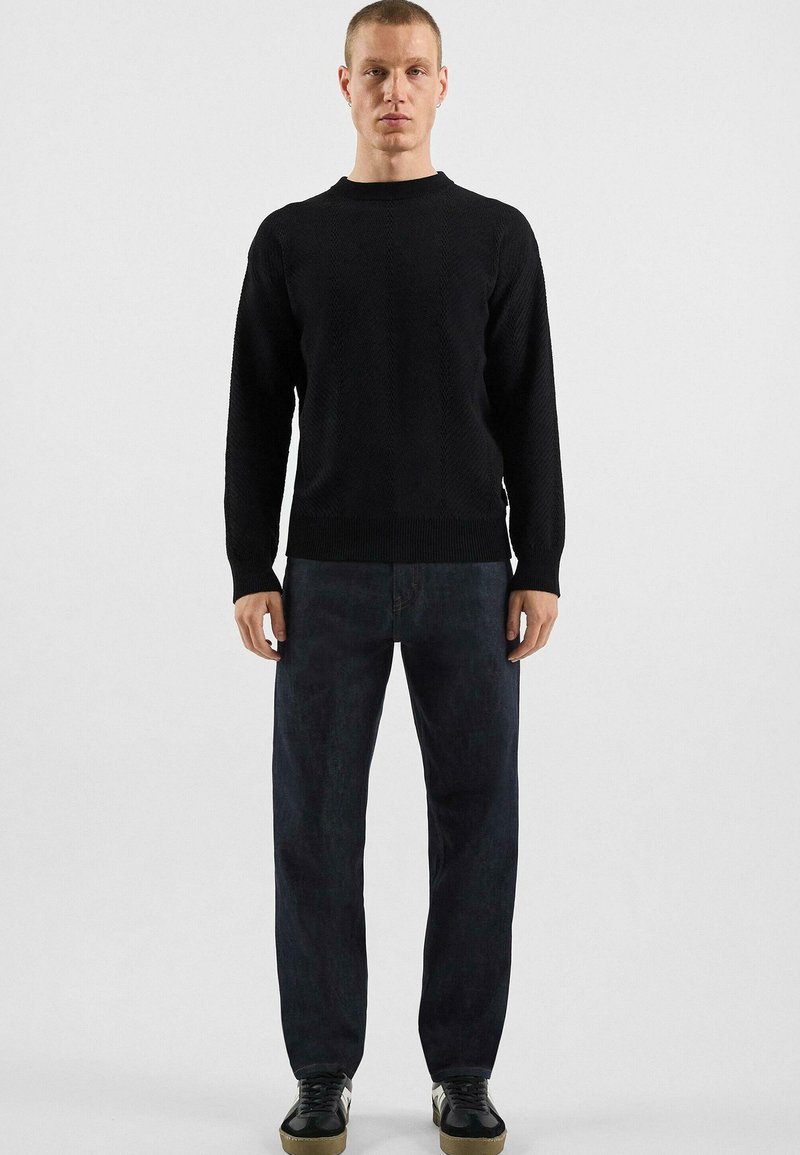 Pull en tricot noir, col rond, manches longues ; associé à un jean droit foncé et des baskets noires avec une semelle texturée.