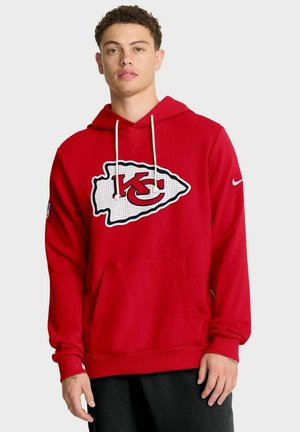 Giovane uomo che indossa un hoodie rosso dei Kansas City Chiefs con lacci bianchi e pantaloni neri, in piedi davanti a uno sfondo chiaro e semplice.