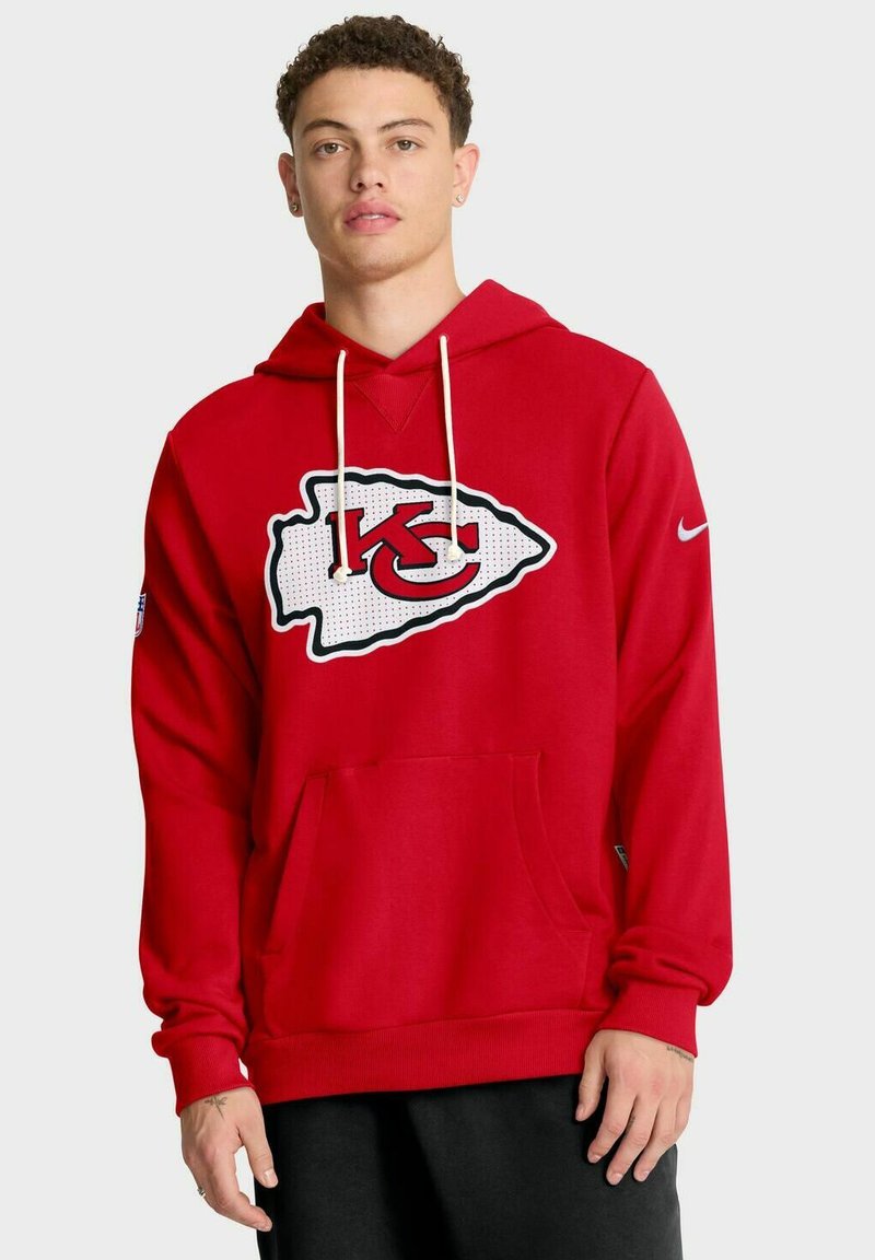 Junger Mann in rotem Kansas City Chiefs Hoodie mit weißen Kordeln und schwarzen Hosen, der vor einem schlichten hellen Hintergrund steht.