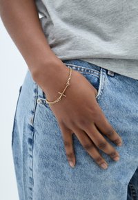 Arany lánc karkötő kereszt charm-mal, egy kézen viselve, amely kék farmernadrágon pihen, a felette látható szürke pólóval.
