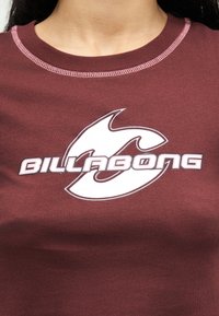 Κοντινό πλάνο ενός ατόμου που φοράει μπορντό πουκάμισο με ροζ ραφές και λευκό λογότυπο Billabong στο κέντρο του στήθους.