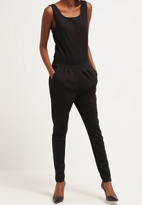 Tuta nera con scollo a barchetta, design senza maniche, tasche laterali, parte anteriore plissettata e gambe affusolate. Materiale liscio e stretch.