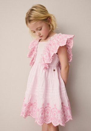 Noor juustega noor tüdruk, kes kannab varrukateta roosat kleiti volangiliste tikitud õlgadega, vaatab alla neutraalse tausta taustal.