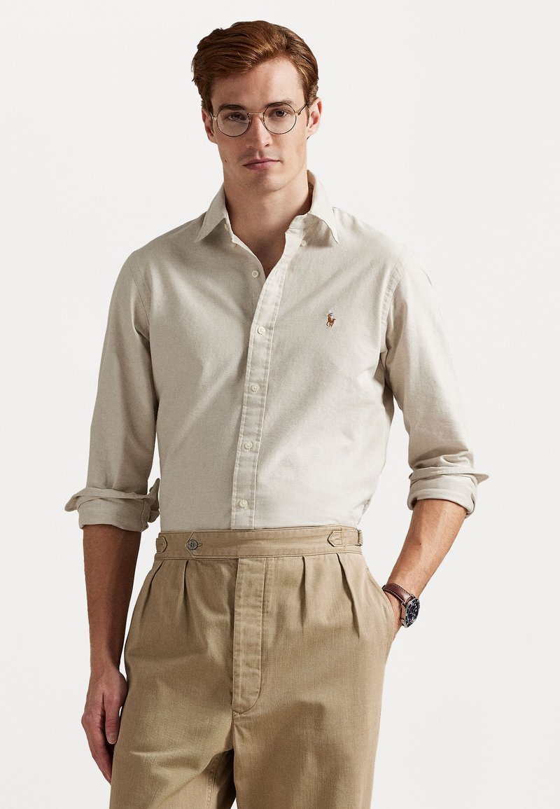 Beige button-up shirt with rolled sleeves, polo logo on the chest, paired with high-waisted tan trousers featuring side pockets and metal accents.